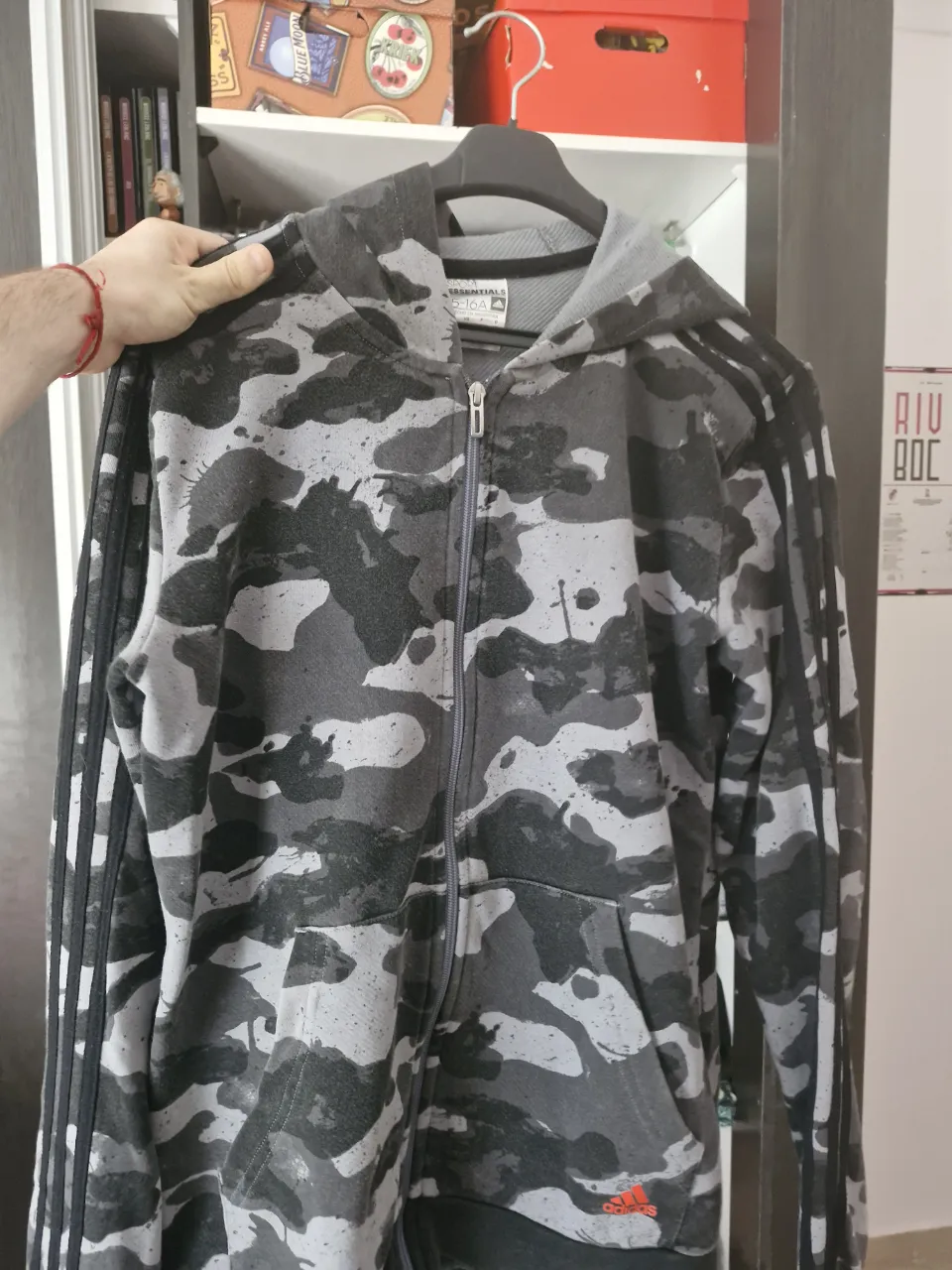 Buzo con capucha y cierre completo, estampado camuflado en tonos grises y negros. Posee bolsillos frontales y las icónicas tres tiras de la marca en las mangas.