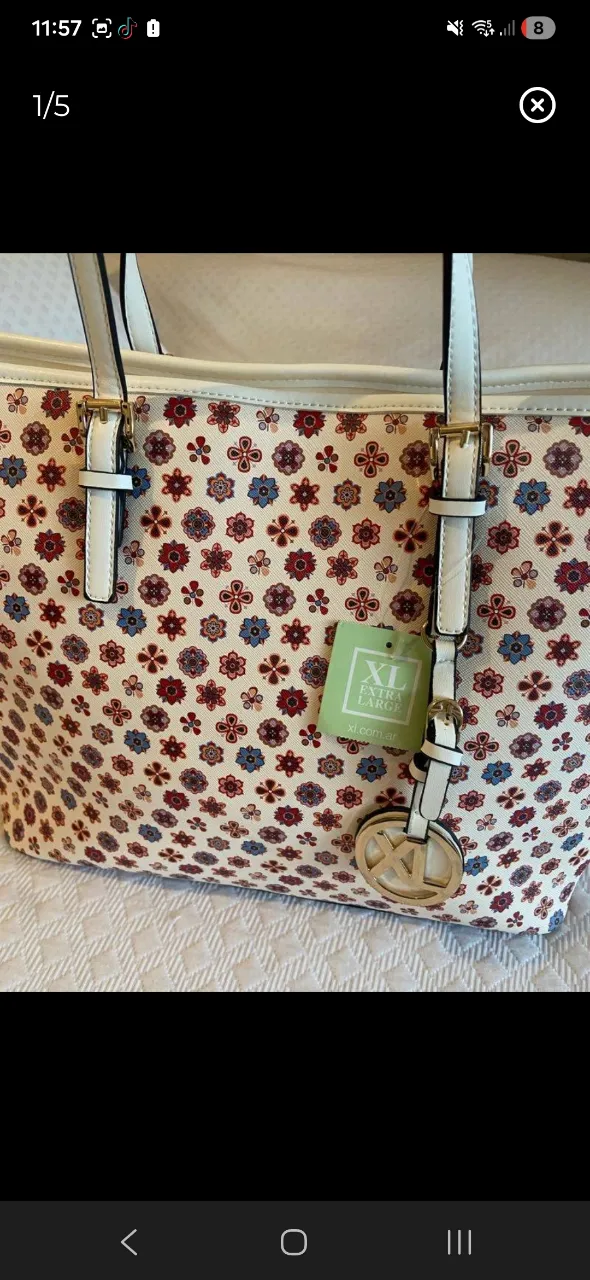 Bolso tote de cuero sintético con un estampado floral vibrante en tonos rojos, azules y blancos sobre una base clara. Posee manijas superiores y un dije metálico dorado con el logo de la marca. El interior es amplio y forrado en rojo, con bolsillos internos para organización. Ideal para el día a día o salidas casuales.