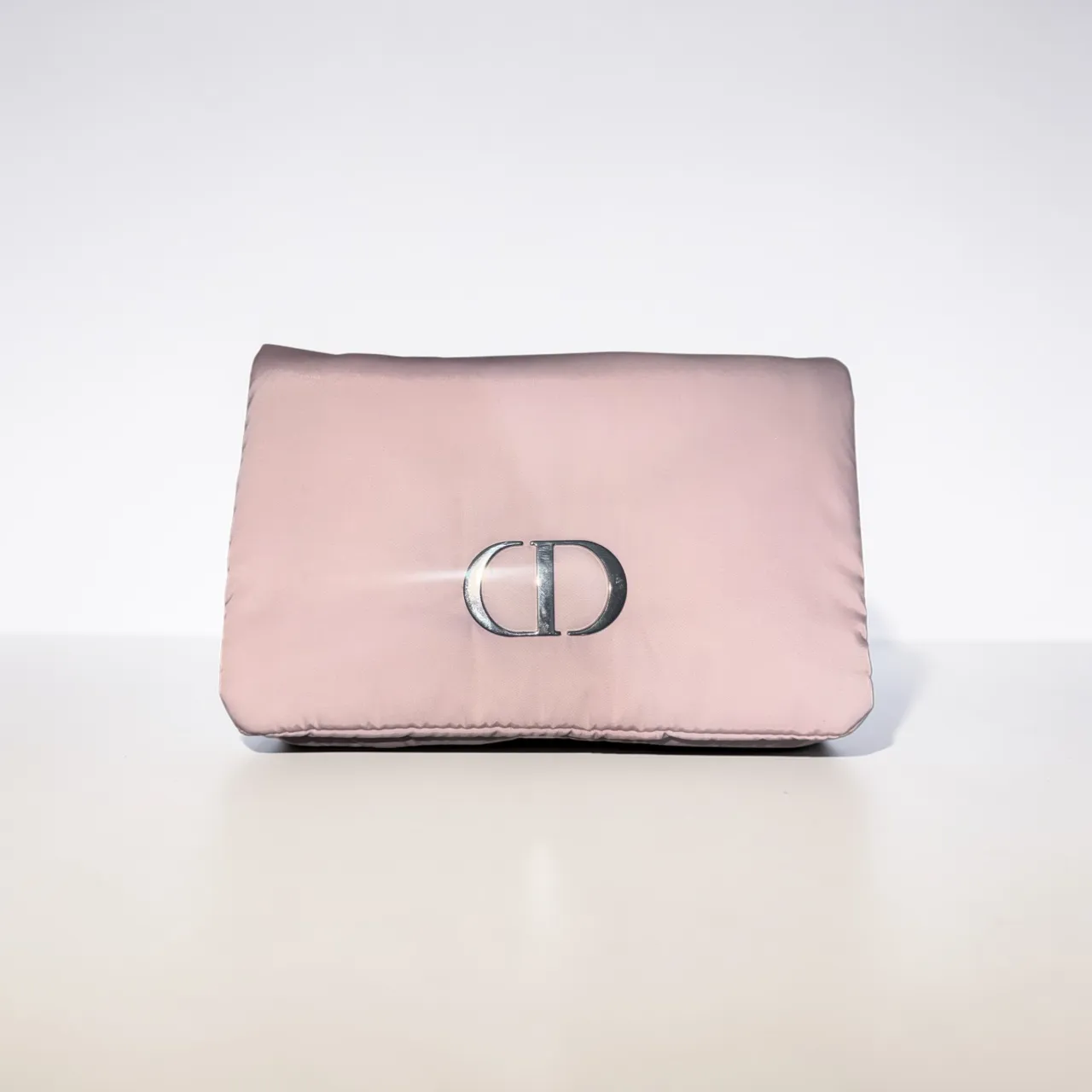 neceser acolchada en color rosa pálido, con el icónico logo 'CD' plateado al frente. Perfecta para llevar tus esenciales con mucho estilo. 
