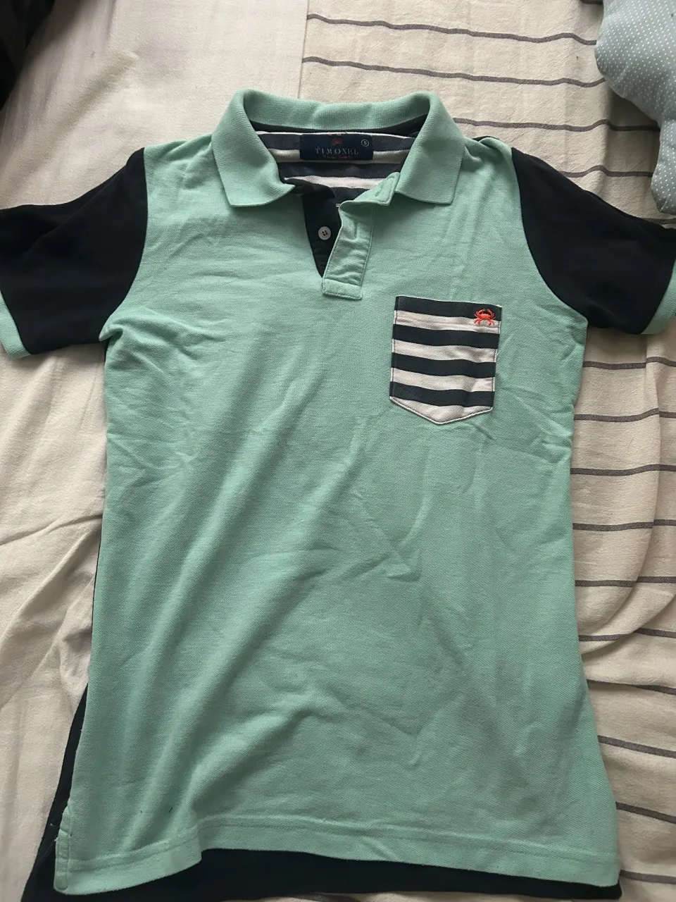 Remera tipo polo de manga corta marca Timonel. La parte delantera es de color verde menta con un bolsillo a rayas blancas y negras. Las mangas y la parte trasera son de color azul marino. El cuello es de color verde menta.