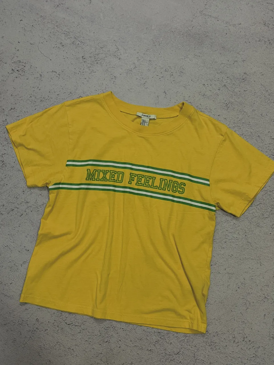 Remera amarilla con estampa "MIXED FEELINGS" en el frente, con dos líneas verdes y una blanca. 