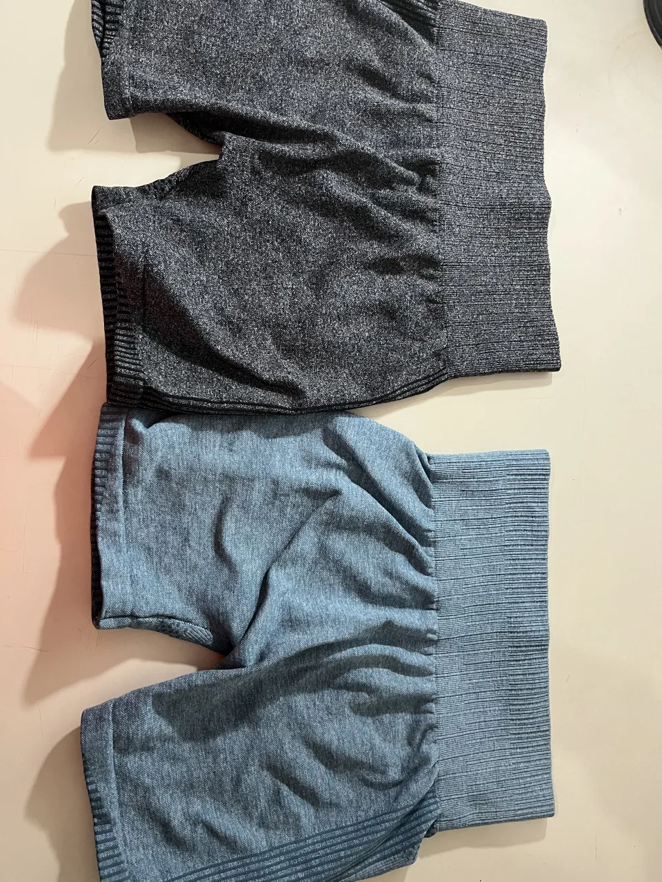 Conjunto de dos shorts deportivos de tiro alto. Uno es gris jaspeado y el otro es azul jaspeado. Ambos tienen cintura ancha y elástica con detalles de tejido acanalado y líneas horizontales en la parte inferior.