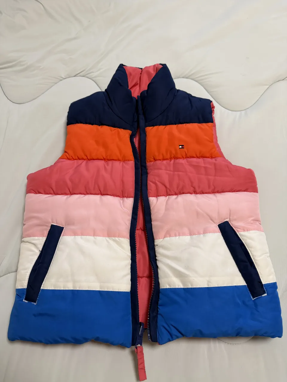 Chaleco acolchado a rayas horizontales en colores azul marino, naranja, rosa, blanco y azul. Tiene cuello alto y cierre al frente. Posee bolsillos laterales con ribetes en azul marino. Reversible.