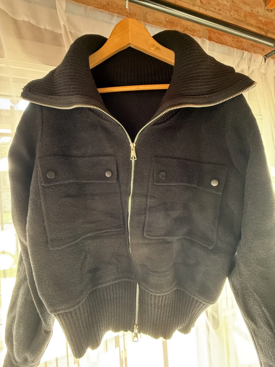 Campera negra con cuello alto tejido, cierre metálico y dos bolsillos frontales con botones a presión. Puños y cintura tejidos.