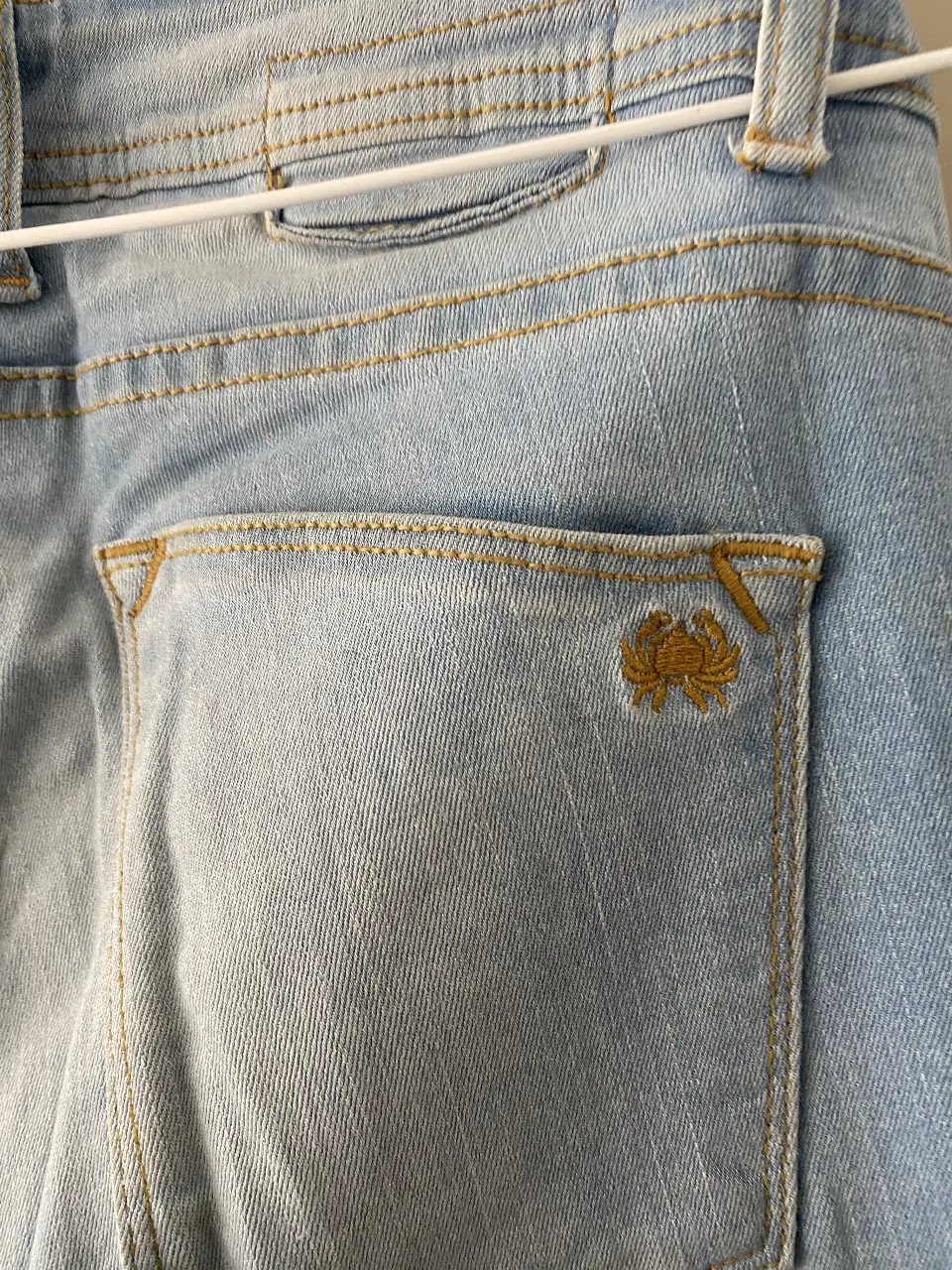Jeans celeste de hombre, corte recto, con detalles de costura en hilo amarillo. Posee un bolsillo trasero con un bordado.
