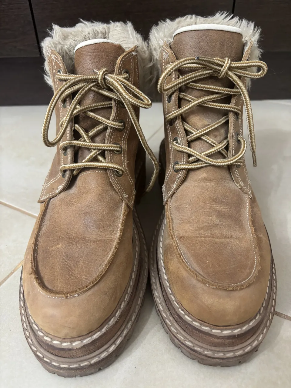 Botas de cuero marrón con cordones y detalles de costura blanca. Tienen un forro interior de pelo sintético color crudo en la parte superior.