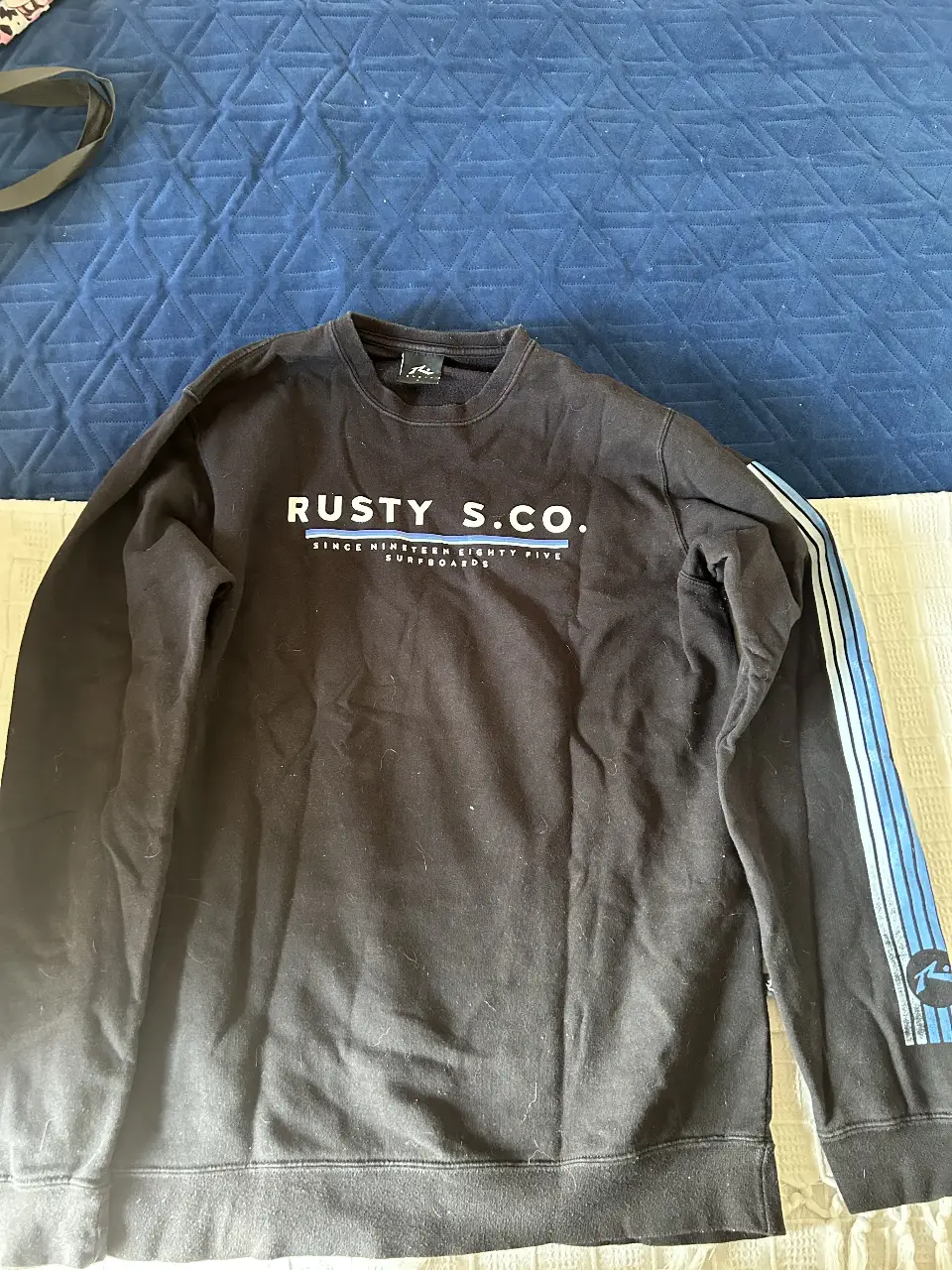 Buzo de cuello redondo negro de la marca Rusty S.Co. con estampado blanco en el pecho y detalles en azul en la manga. Ideal para un look casual y cómodo.