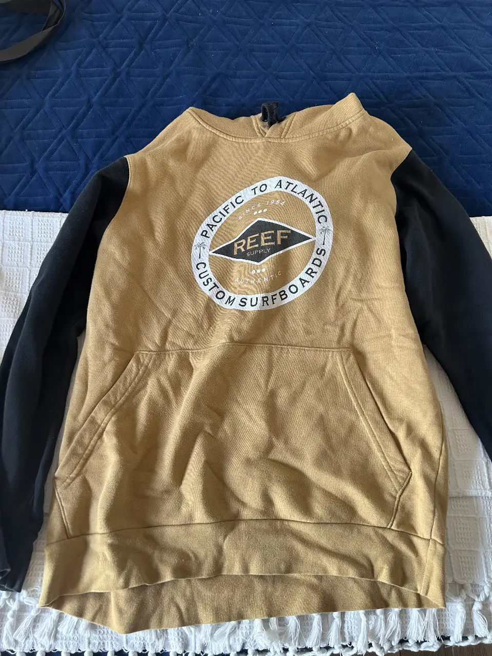 Buzo canguro Reef para hombre, color mostaza con mangas negras. Con capucha y bolsillo frontal. Estampado en el pecho con el logo de la marca.