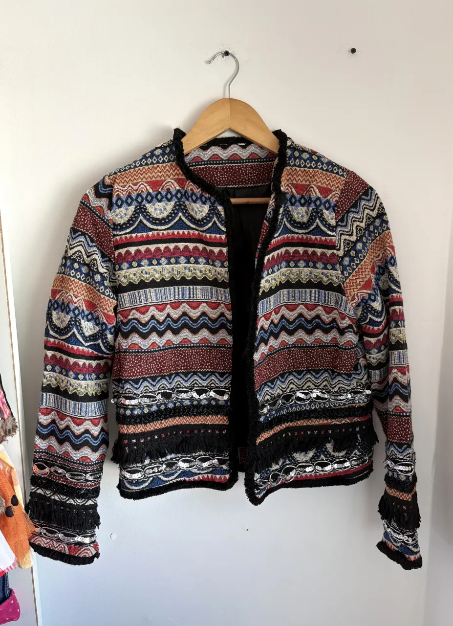 Campera corta con diseño de franjas y figuras geométricas en varios colores. Posee detalles de flecos en los puños y en el borde inferior. Cuello redondo y sin botones.
Es para media estación/invierno