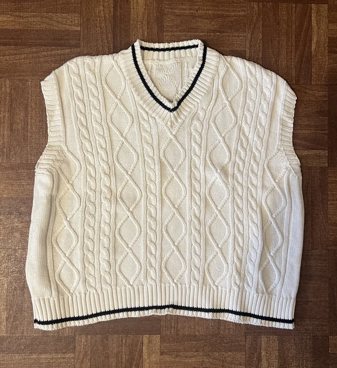 Sweater tejido sin mangas, color blanco roto con detalles en negro. Posee un diseño de trenzas y rombos en el tejido, con cuello V y puños y cintura elastizados con rayas negras. Ideal para un look casual y canchero.