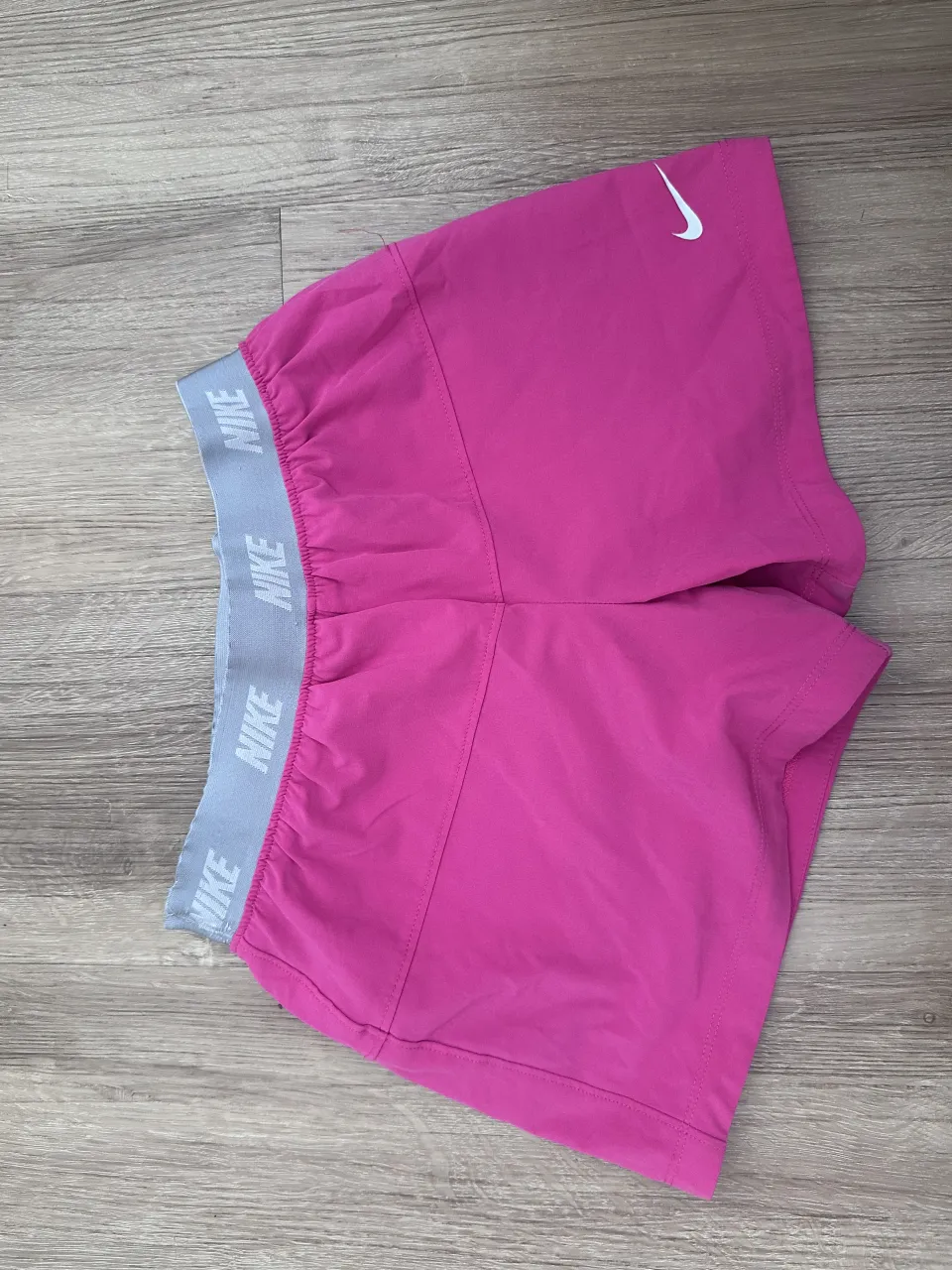 Shorts deportivos color fucsia con cintura elastizada gris con logo NIKE repetido. Logo NIKE blanco en la parte inferior derecha.