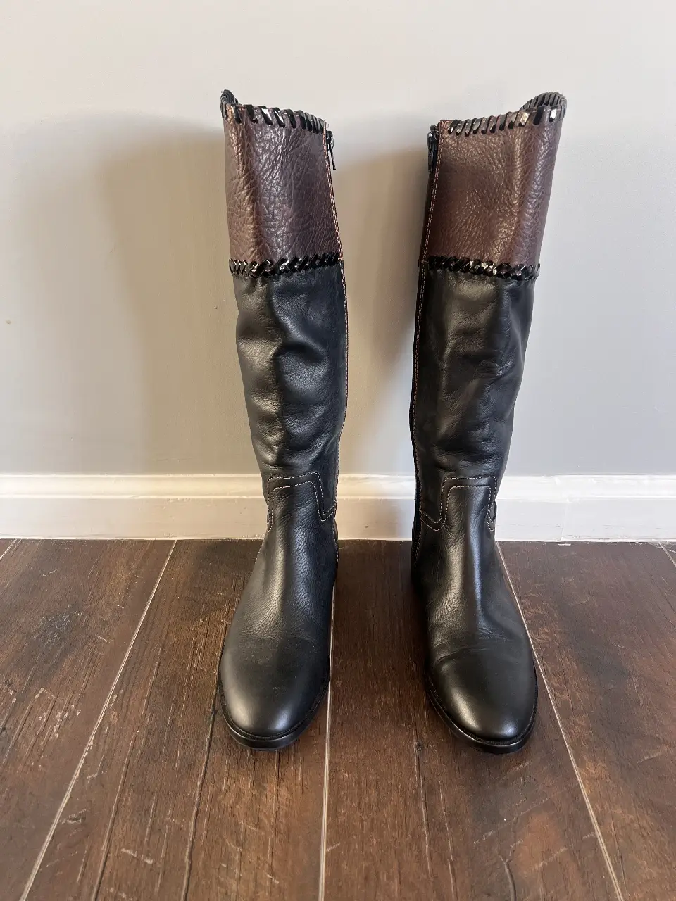 Botas de caña alta para mujer, combinando cuero negro en la parte inferior con un detalle en cuero marrón en la parte superior. Diseño elegante con detalles de costura y cierre lateral. Ideales para un look sofisticado y moderno.