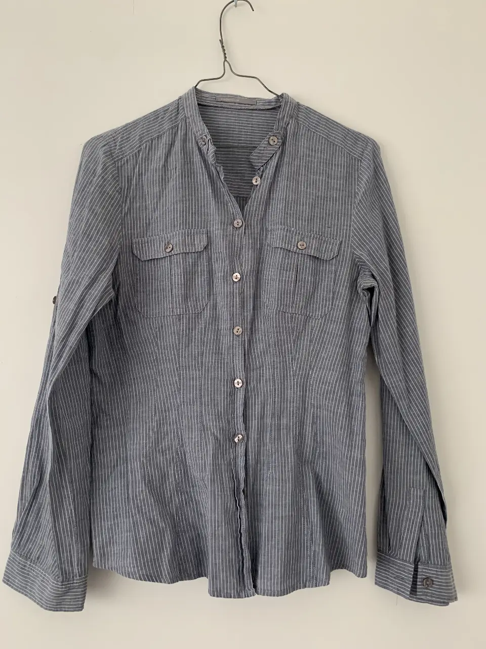 Camisa de mujer a rayas verticales en tonos grises y blancos. Diseño clásico con cuello mao, bolsillos delanteros y mangas largas con detalle de botón en los puños. Ideal para un look casual y versátil.