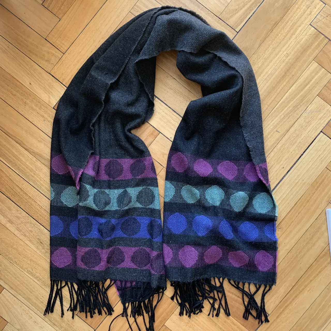 Bufanda de lana con diseño de círculos en tonos violeta, azul y verde sobre fondo gris oscuro. Ideal para abrigar y darle un toque de color a tus outfits de invierno. marca Fallabella 