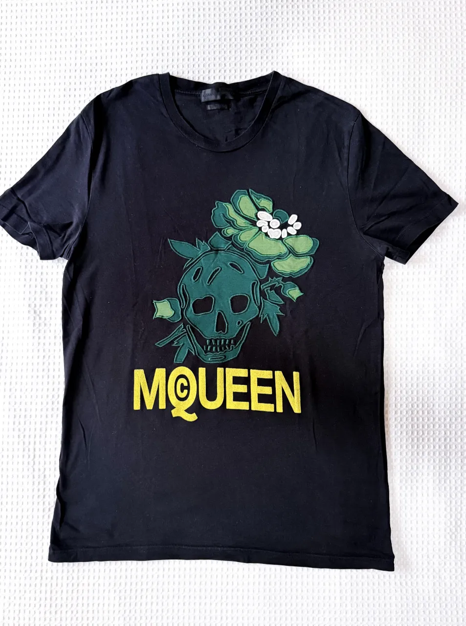 Remera Alexander McQueen negra de manga corta con estampa de calavera verde y flores, y logo "MCQ" en amarillo, con detalle de aplique blanco a cada lado con la calavera bordada. Talle L. 
Medida de sisa: 50 cm.