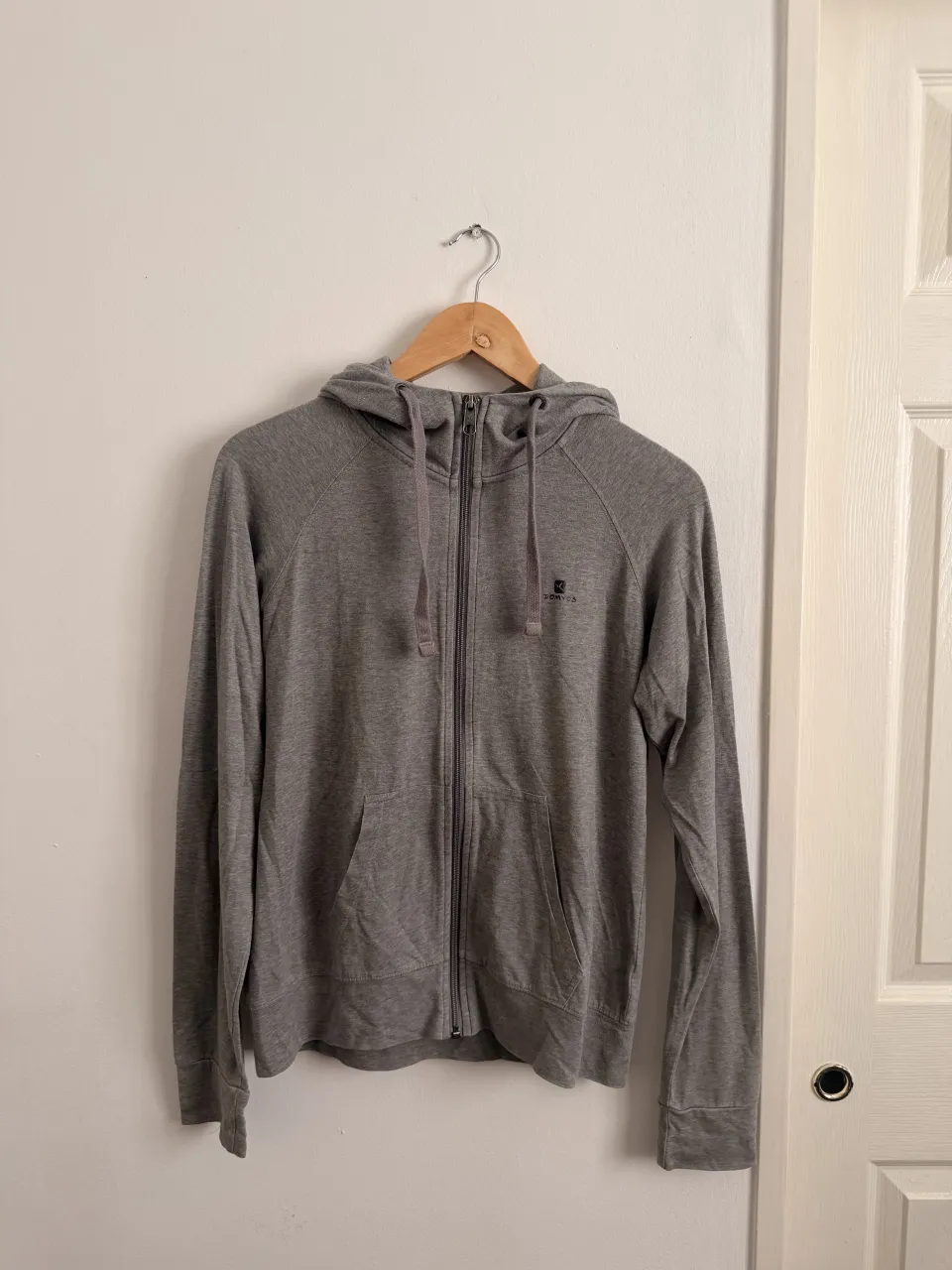 Buzo gris (DECATHLON DOMYOS) con capucha y cierre completo. Posee dos bolsillos frontales tipo canguro y cordones para ajustar la capucha.