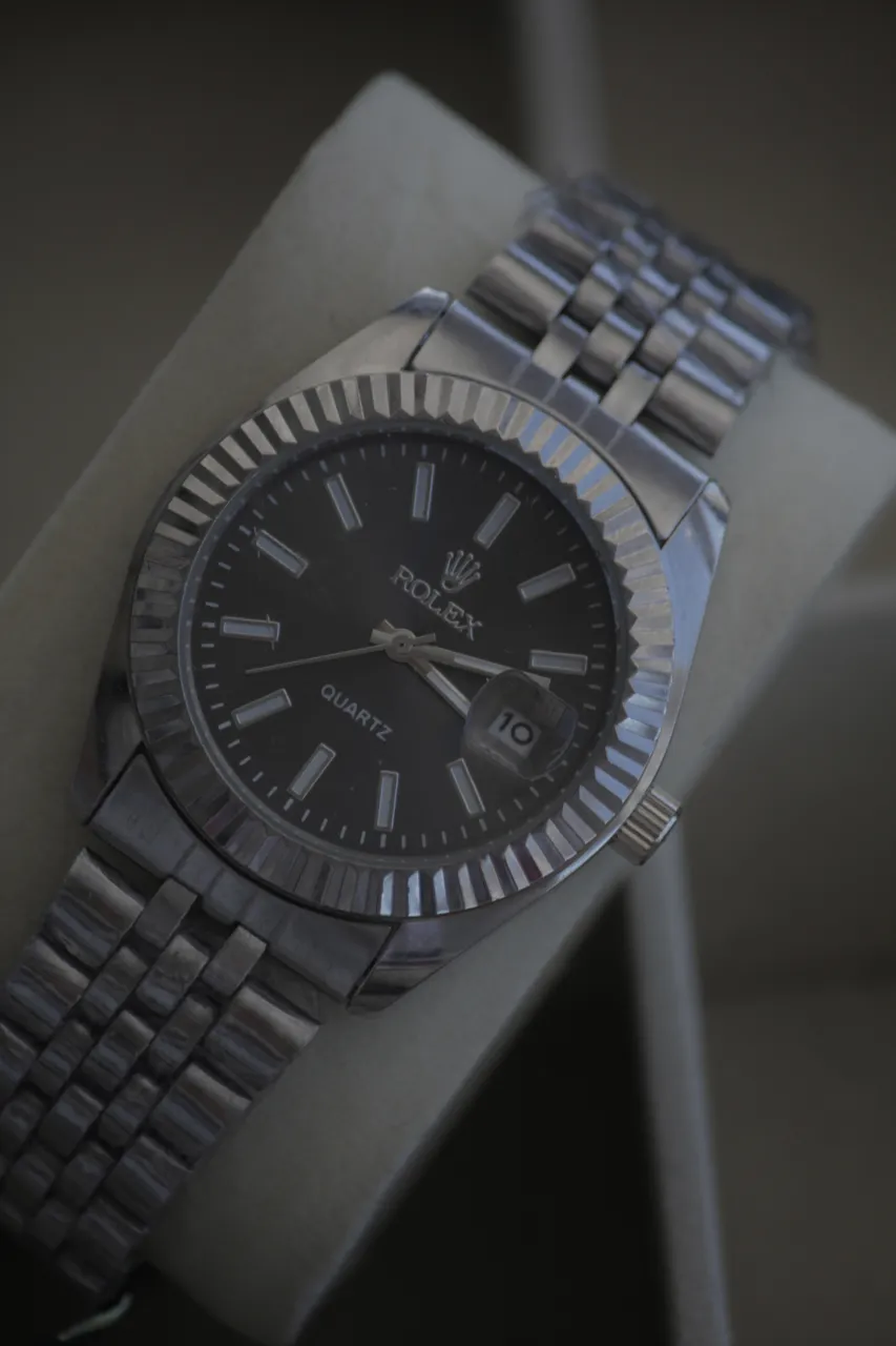 Reloj de pulsera con malla metálica plateada y cuadrante oscuro. Posee detalles de cronógrafo y la marca visible en el cuadrante.