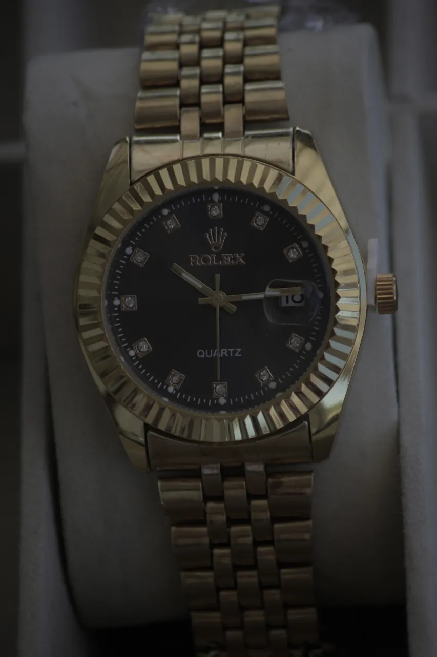 Reloj de pulsera dorado con malla metálica y cuadrante negro. Presenta detalles de strass en los marcadores de hora y un pequeño visor para la fecha.