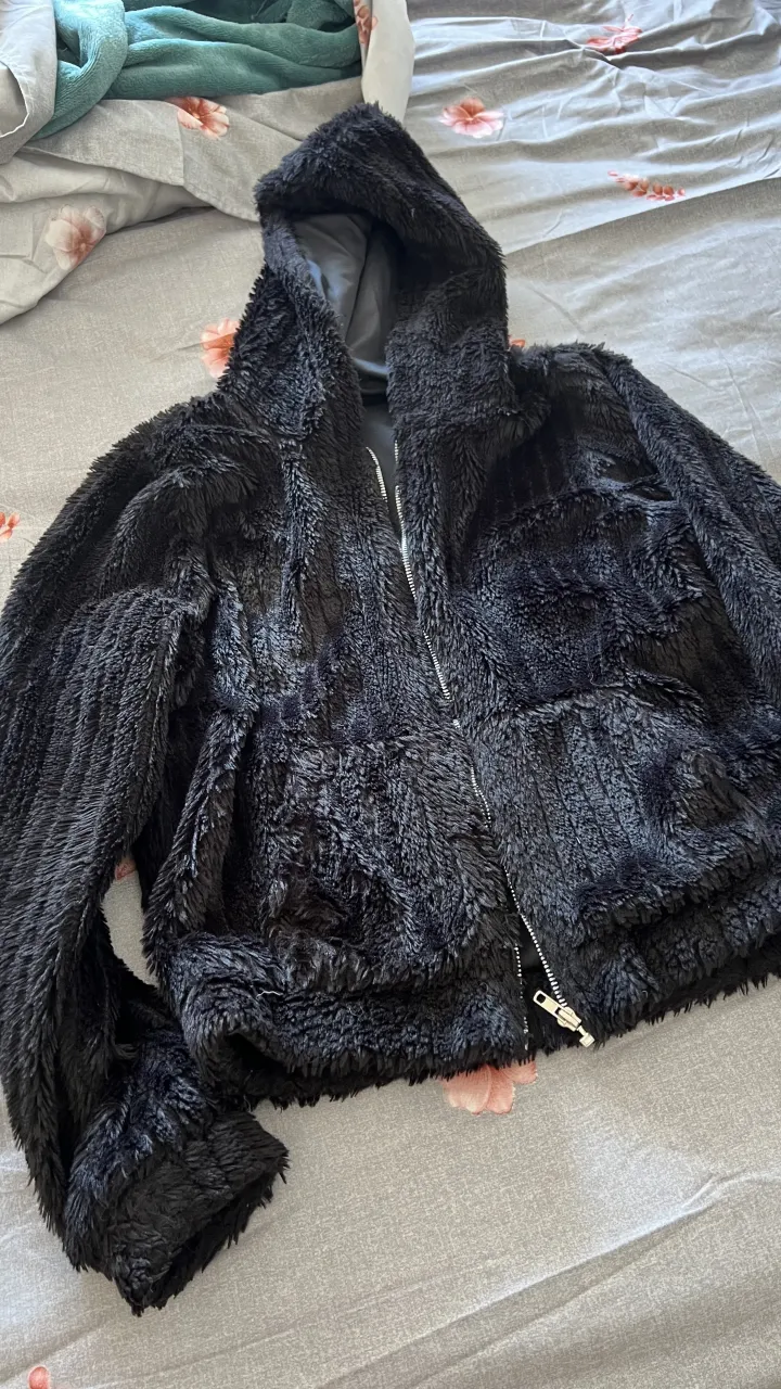 Abrigo de peluche negro con capucha y cierre. Súper suave y calentito, ideal para los días fríos. Tiene bolsillos al frente y un diseño moderno.