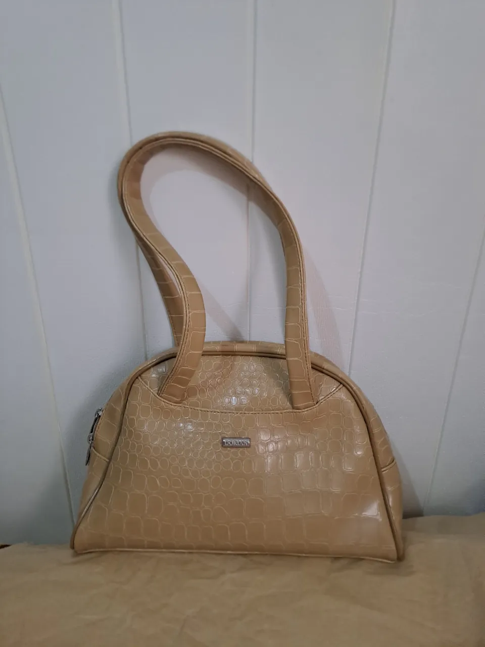 cartera de mano color beige con textura de cocodrilo. Posee dos manijas cortas y un cierre superior. Incluye un detalle metálico con el nombre de la marca.