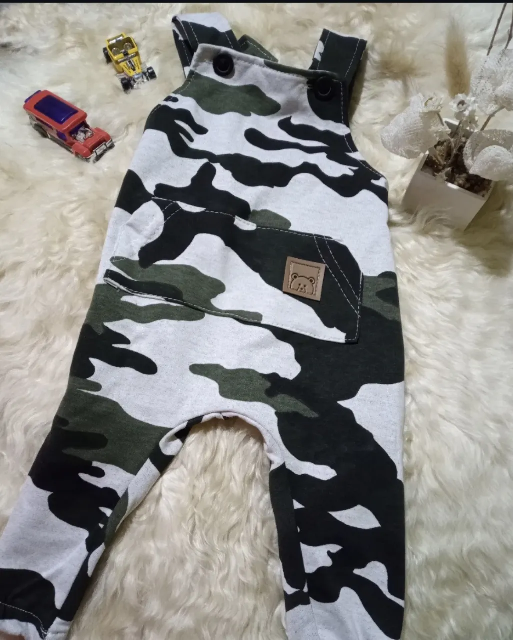 Enterito de bebé con estampado camuflado en tonos verde, blanco y negro. Posee dos botones en los breteles y un bolsillo superior con un aplique de oso.