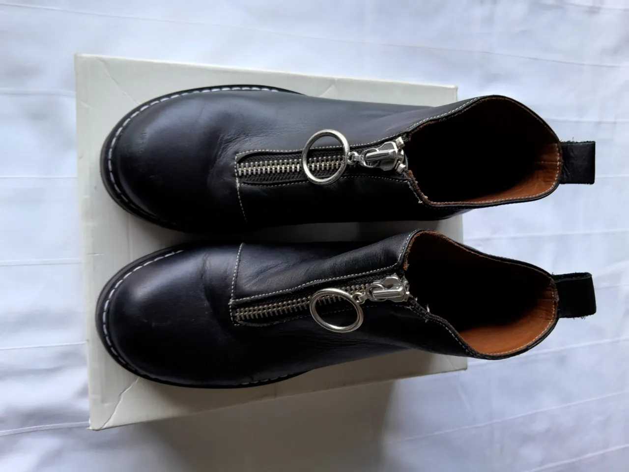 Botas negras de cuero con cierre metálico al frente y detalle de aro. Costuras blancas visibles en el borde superior y puntera. Suela de goma negra con relieve.