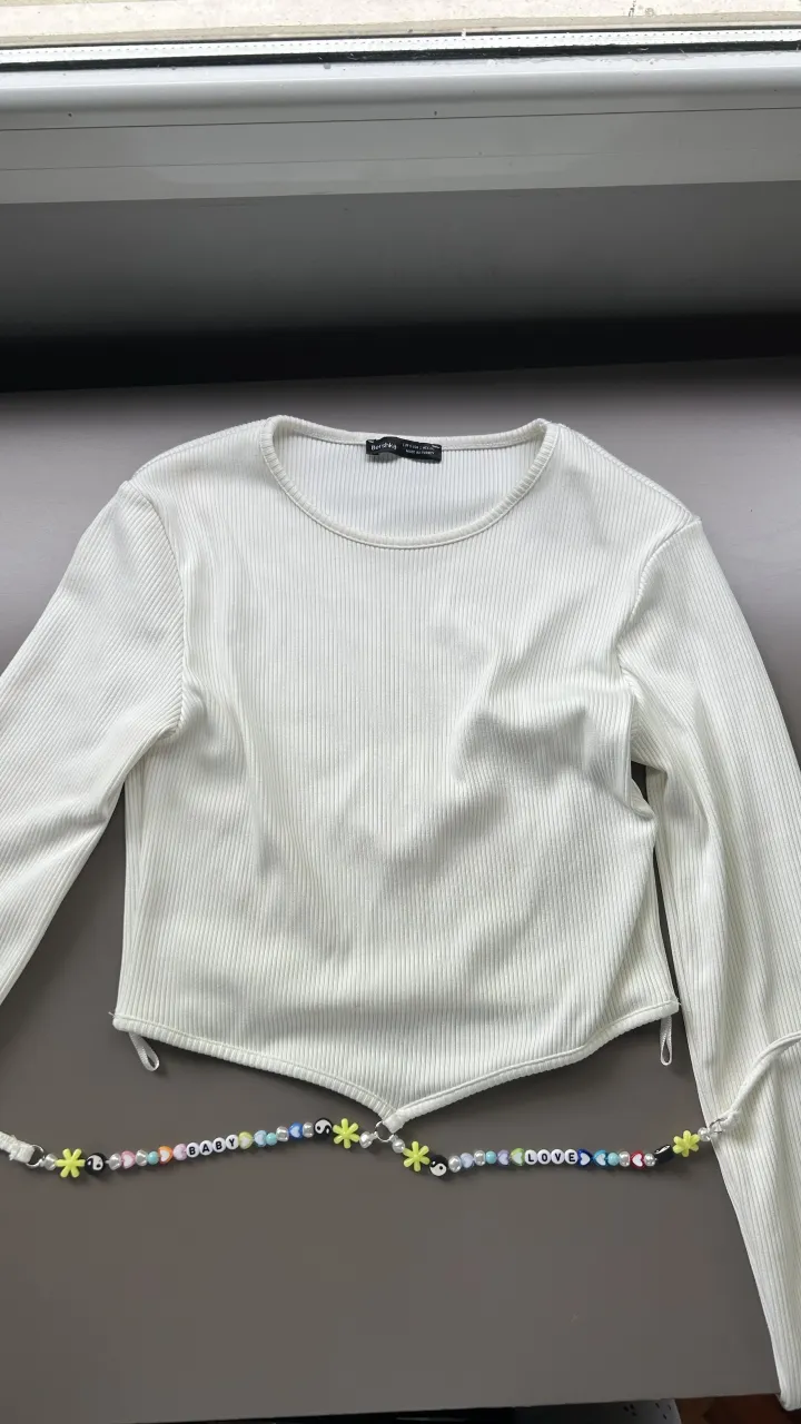 Remera blanca de manga larga con cuello redondo y detalles acanalados. Posee un accesorio de cuentas de colores y dijes colgando de la parte inferior.