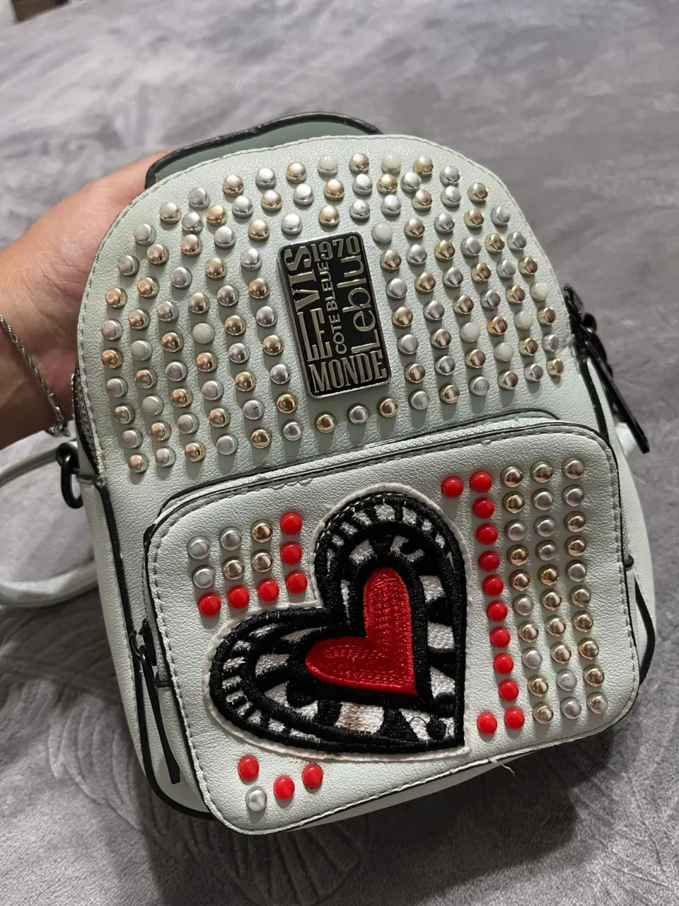 Mini mochila blanca con tachas plateadas y doradas. Posee un aplique de corazón rojo bordado con detalles en negro y tachas rojas.