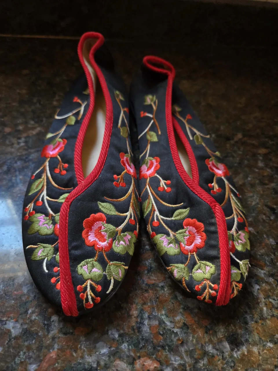 Zapatiros bajas de tela negra con bordados florales en rojo, rosa y verde. Tienen un ribete rojo alrededor de la abertura y la punta. Son originales de China. Hirma chica.