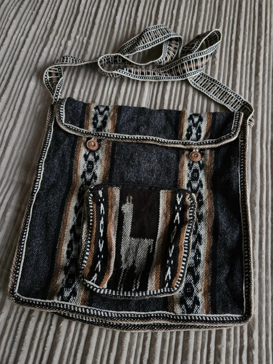 Bolso de tela con correa tejida y detalles de diseño étnico. Posee un bolsillo frontal con cierre y dos botones de madera para el cierre principal.