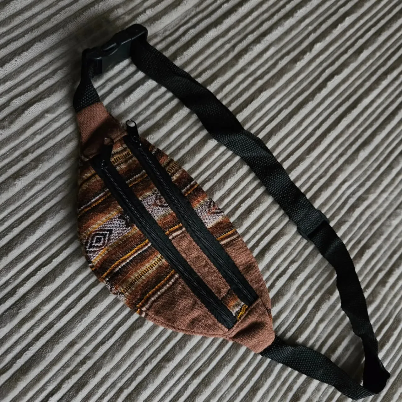 Riñonera de tela con diseño étnico en tonos marrones, ocres y blancos. Posee tres cierres frontales y una correa ajustable con hebilla plástica.