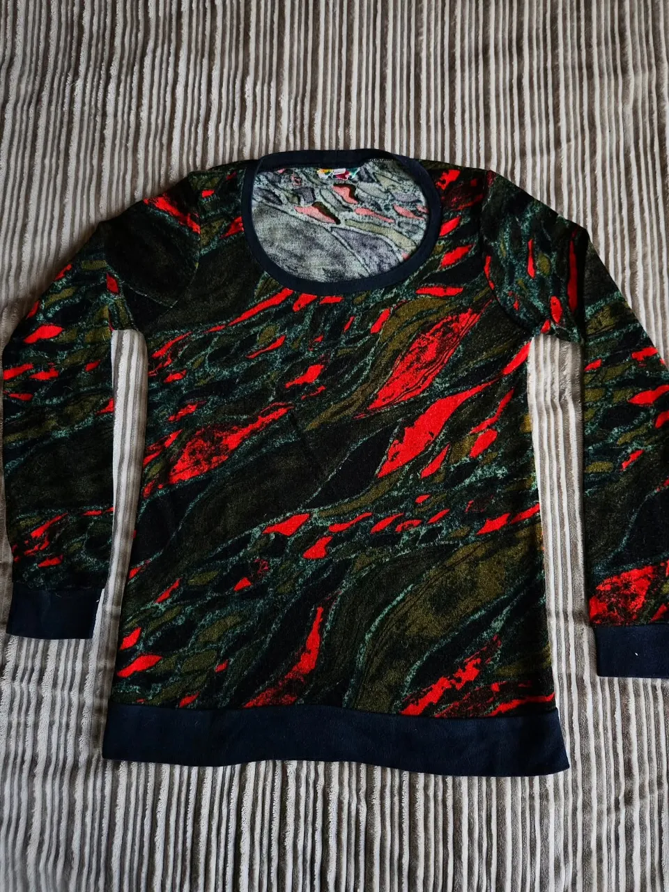 Buzo de lanilla de manga larga con estampa abstracta en tonos verdes, negros y rojos. Cuello redondo y puños en negro.