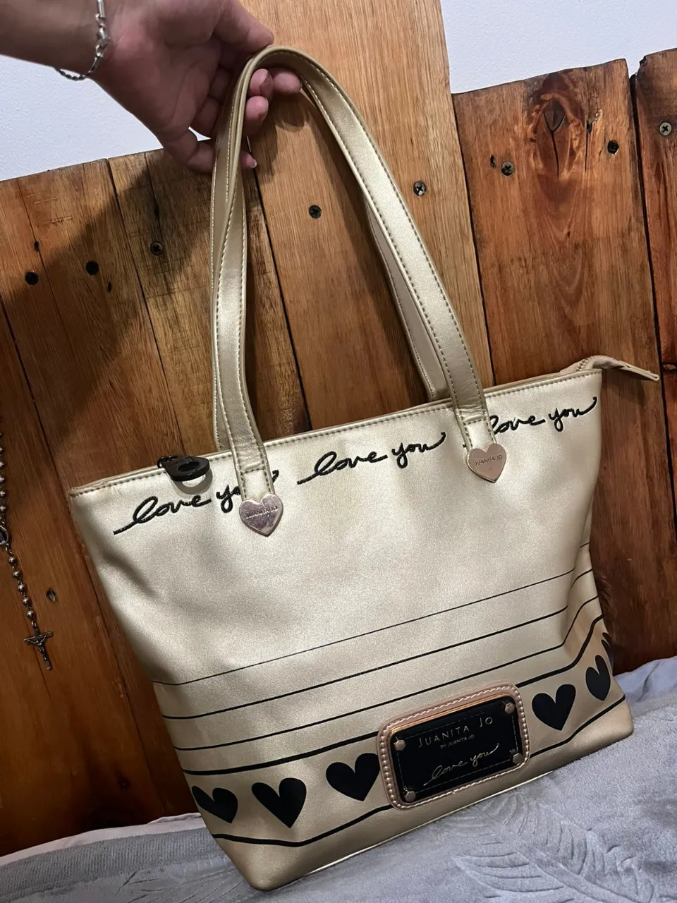 Bolso tote color dorado con inscripciones "love you" en negro y detalles de corazones negros. Posee un bolsillo exterior con placa metálica negra y dorada con el logo "JUANITA JO".