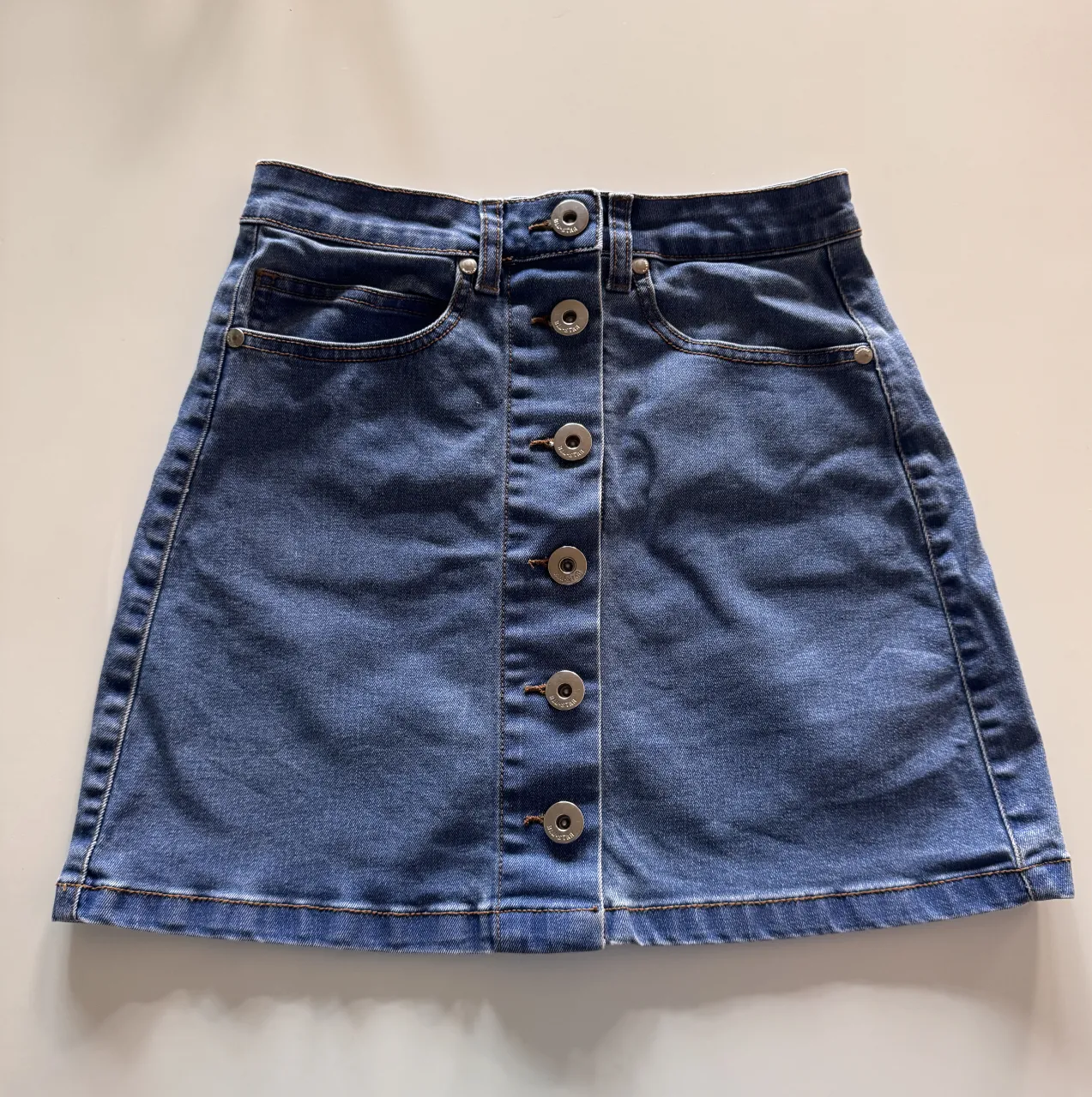 Pollera de jean tiro alto, con botones al frente y bolsillos. Un clásico que no puede faltar en tu guardarropa, ideal para combinar con tus tops favoritos (cintura: 30 cm ancho)